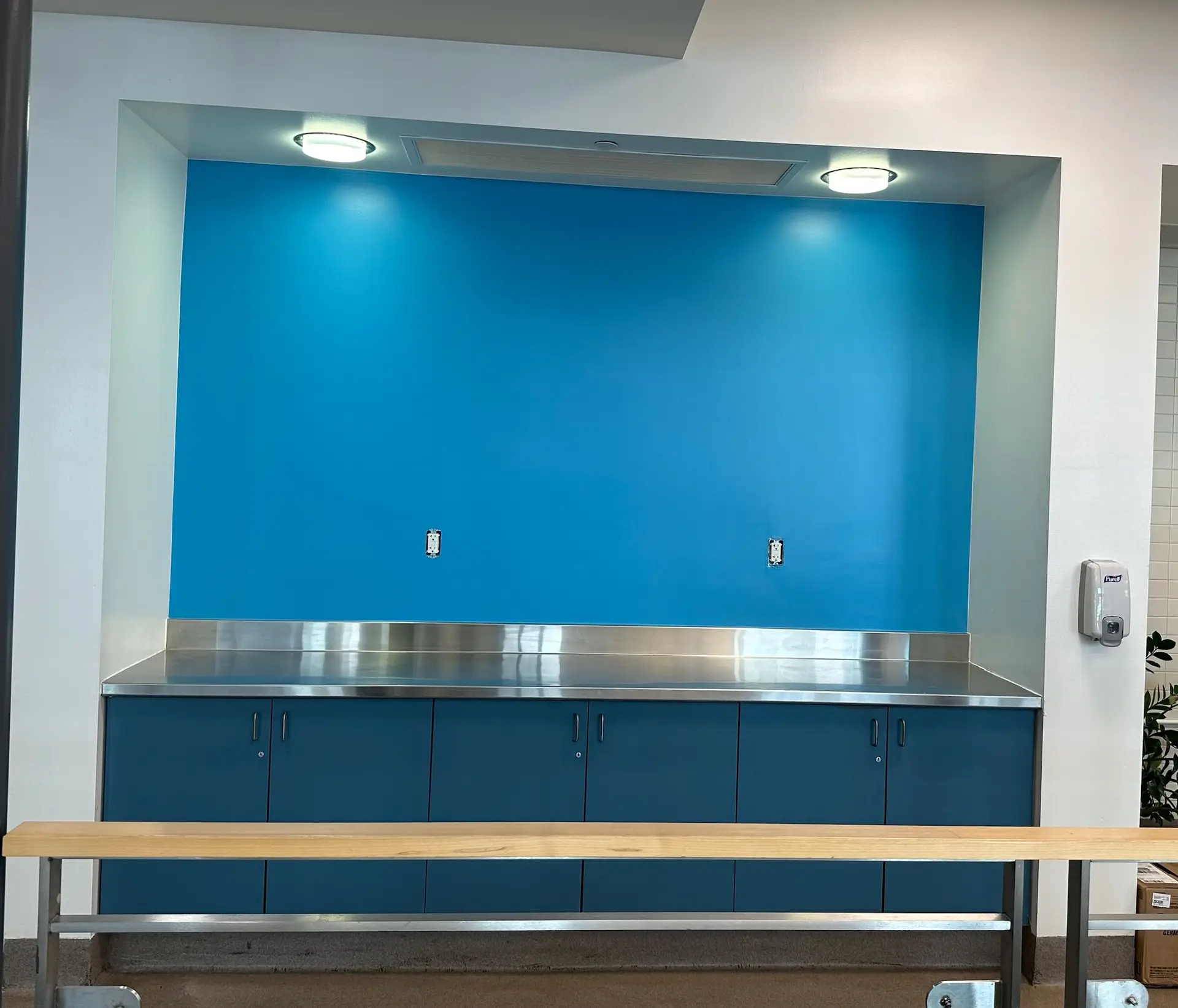 A built-in counter area featuring dark blue lower cabinets and a sleek stainless steel countertop. The backdrop is a vibrant light blue accent wall with two electrical outlets, illuminated by overhead ceiling lights in a recessed alcove.