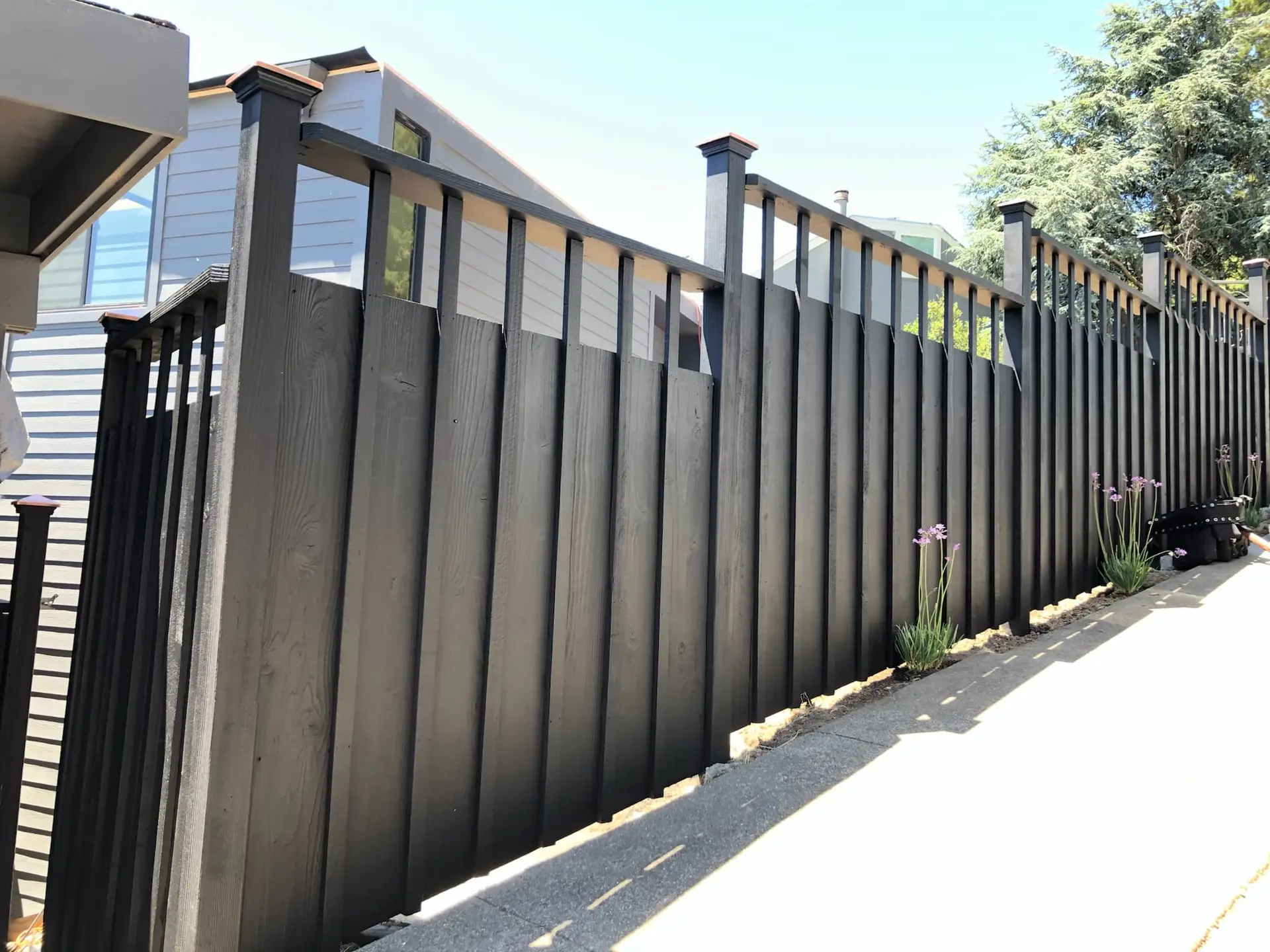 A stylish matte black wooden privacy fence built on an incline next to a concrete path. The fence features vertical planks with unique open vertical slats at the top, supported by sturdy posts with caps. Small purple flowers are growing along the base, with houses and trees in the background.